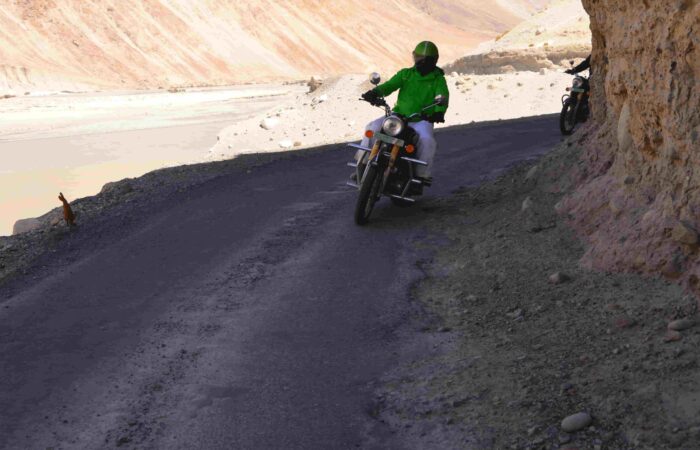 Riders on Royal Enfield motorcycles crossing a mountain bridge in the foothills of the Himalayas during Into Wild Himalaya’s guided adventure tour in North India.