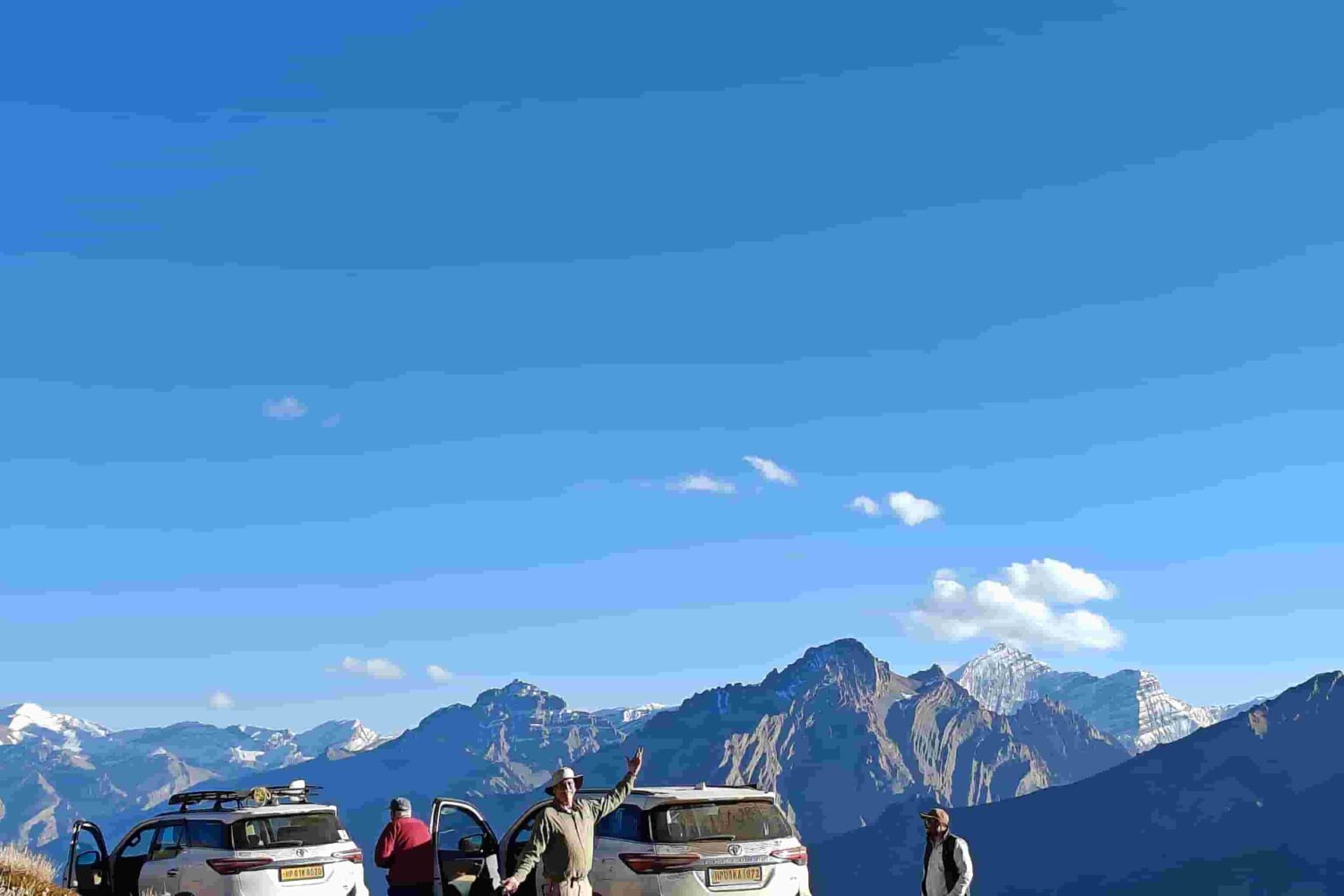 Travellers standing beside white 4x4 vehicles on a high-altitude Himalayan dirt road with rugged mountains, clear sky, and scenic views during a guided road trip.