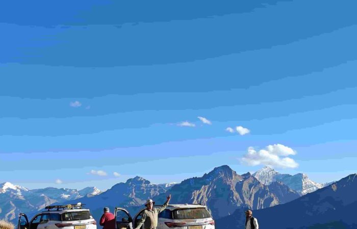 Travellers standing beside white 4x4 vehicles on a high-altitude Himalayan dirt road with rugged mountains, clear sky, and scenic views during a guided road trip.