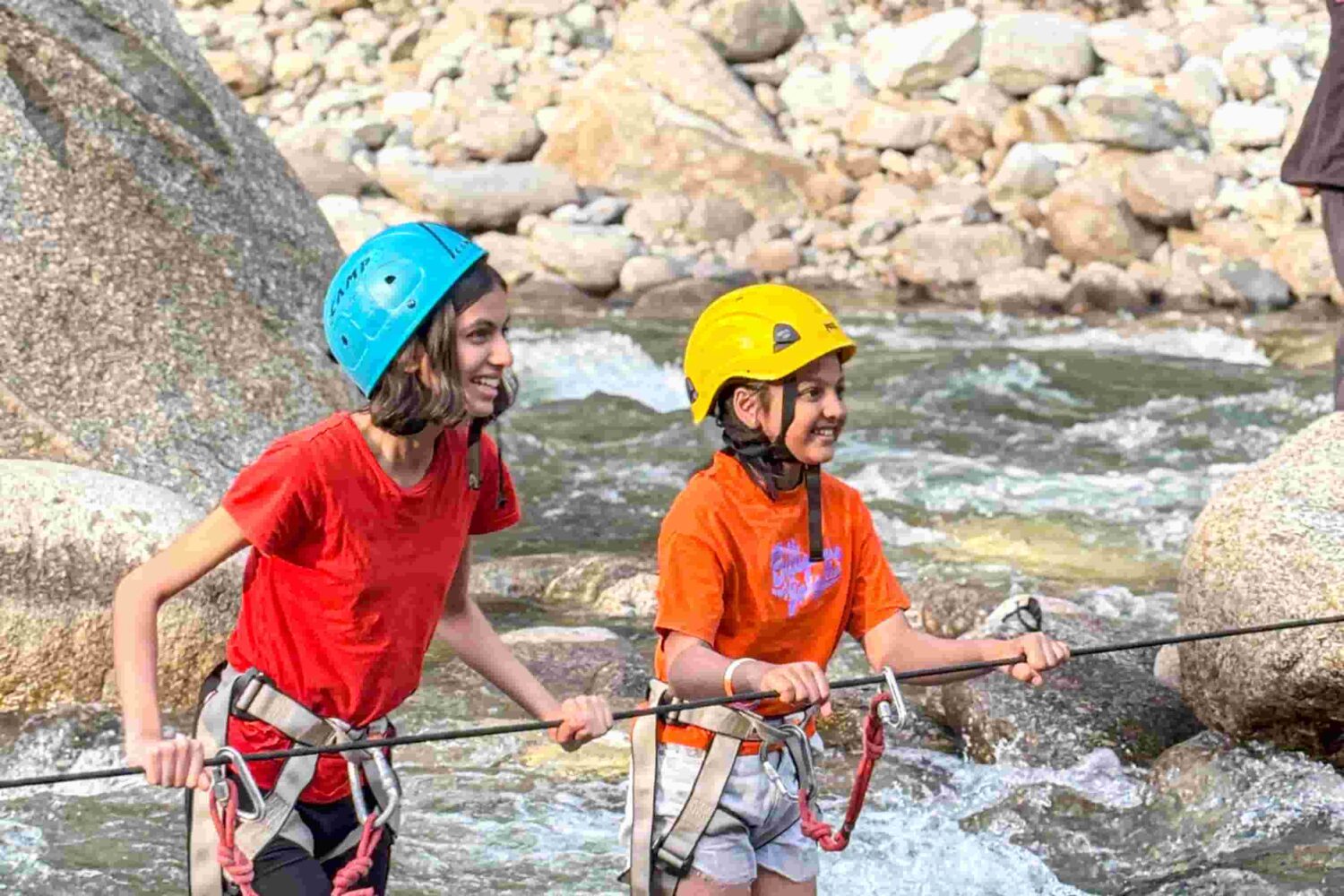 Two participants crossing a river using a safety rope during a weekend camping adventure in Tirthan Valley, India.