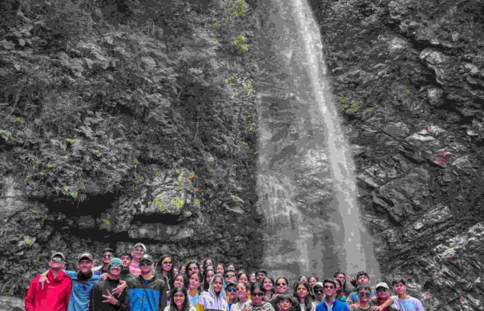A group of campers posing in front of a tall waterfall during a weekend camping trip in Tirthan Valley, India.