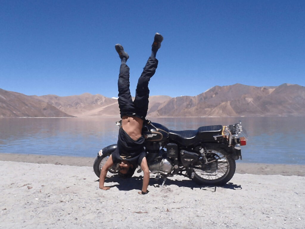 This image represents the spirit of Into Wild Himalaya’s motorcycle expeditions adventure blended with mindfulness and connection to nature.
The rider performing a handstand beside a mountain lake symbolizes balance, freedom, and harmony with the Himalayas core values behind IWH’s expeditions.