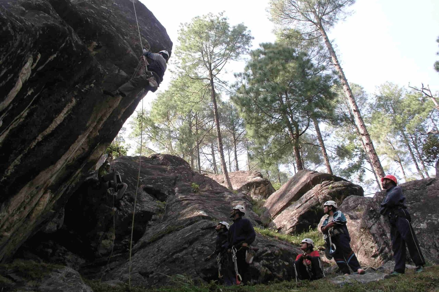 Trainee practicing natural rock climbing during the Private Basic Mountaineering Course in Manali India while the instructor team supervises in the forest training zone.