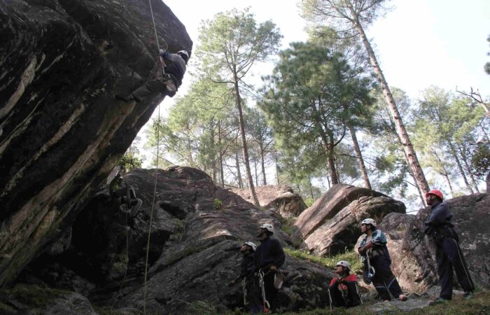 Trainee practicing natural rock climbing during the Private Basic Mountaineering Course in Manali India while the instructor team supervises in the forest training zone.