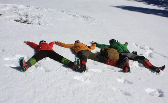 Three trekkers lying on fresh snow with arms and legs spread wide, relaxing under the Himalayan sky during the Patalsu Peak Day Hike near Manali.