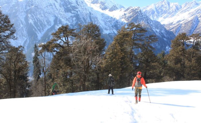Two trekkers walking down a snow-covered meadow during the Patalsu Peak Day Hike near Manali, with the majestic Dhauladhar range shining in the background.