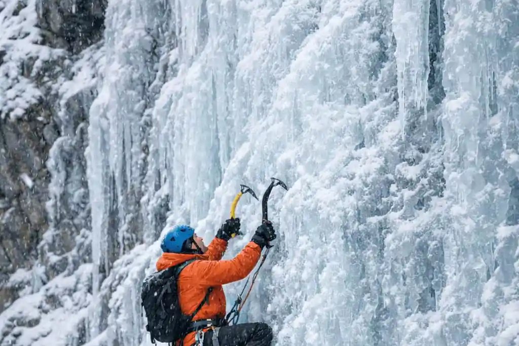 Climber in an orange jacket swinging an ice axe on a frozen blue waterfall in the Indian Himalayas